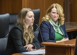 Emily Myatt sits at the testifiers' table and speaks into a microphone to someone out of view. Representative Halverson sits next to her and watches as she speaks.